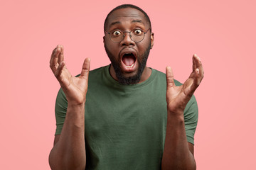 Impressed surprised black guy opens mouth widely, holds plams near face, feels shocked and amazed, expresses disbelief, gestues actively, poses on pink studio background. Facial expressions, emotions