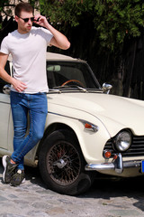 Portrait of a handsome smiling man with sunglasses, male model posing in urban city street next his car. Stylish young guy is standing near white retro car. Classic car, retro car and vehicle concept.