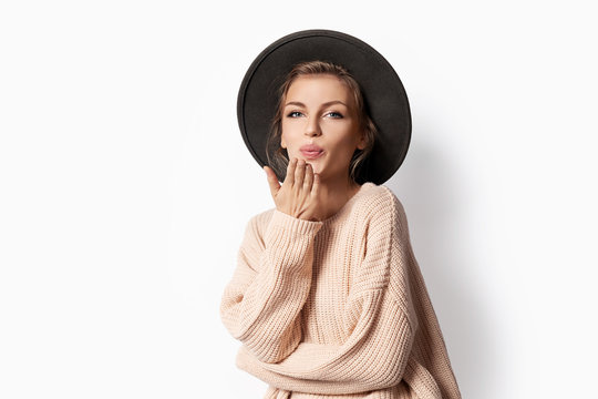 Portrait of  young woman sending air kisses on white background. Indoor photo of beautiful girl in hat posing in studio. Modern fashion concept.