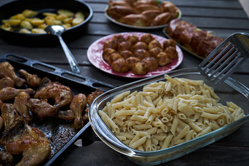 Party snack food table with baked potatoes, fried chicken drumsticks, italian pasta and puff pastry buns bread, close-up. Table with variety food top view. 