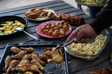 Catering buffet food table with baked potatoes, fried chicken legs, pasta and vegetable salad. People at a party taking different food, outdoor. Colleagues Buffet Party Brunch Dining Concept
