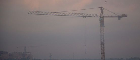 very high construction crane in the morning fog, against the gray sky background