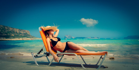 Young redhead girl in black bikini and with hat lying down on lounger on Balos beach, west Crete, Greece. Summertime season vacation, July