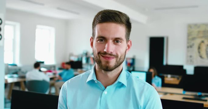 Portrait of young adult mid 30s handsome Caucasian male smiling and looking into camera, busy office in the background. 4K UHD 60 FPS SLOW MOTION