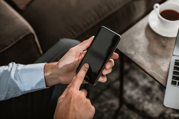 Close up of male hands holding mobile phone. Guy is sitting at coffee table with laptop and hot tea