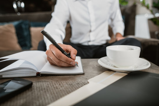 Close up of male hand handling pen and writing down notes in sketchbook. He is having mug with hot drink and using phone while doing some business in informal atmosphere