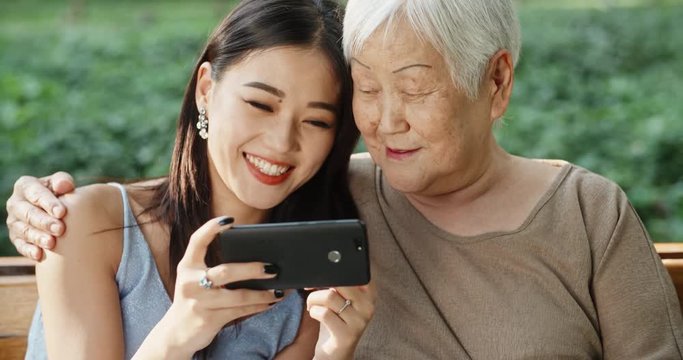 Happy asian Mother and daughter sitting on bench in garden, looking through photos on cellphone, and smiling. closeup 4k