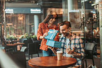 Portrait of positive female showing present for beaming male comrade. He looking at it while telling with cheerful girl indoor. He gesticulating hands during communication