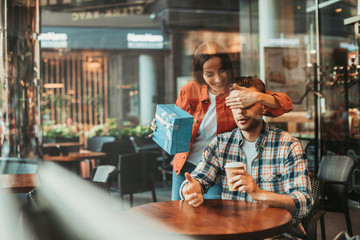 Who is it. Portrait of optimistic woman closing eyes of astonished man while keeping gift for him in arm. He drinking mug of appetizing liquid at desk in cafe