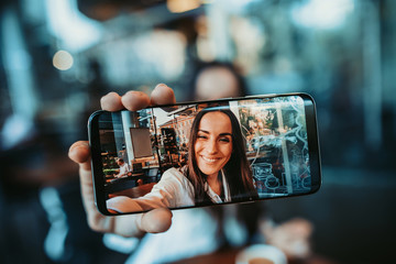 Portrait of beaming girl looking at mobile while taking selfie on it in cafe. Close-up female hand...