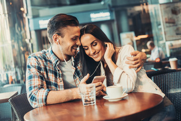 Positive boyfriend embracing outgoing woman while listening music in headset. They situating at table and looking at mobile together