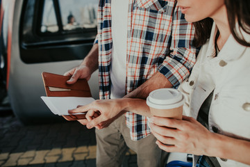 Woman holding cup of delicious beverage in hand while touching tickets for train. She situating near male comrade outside