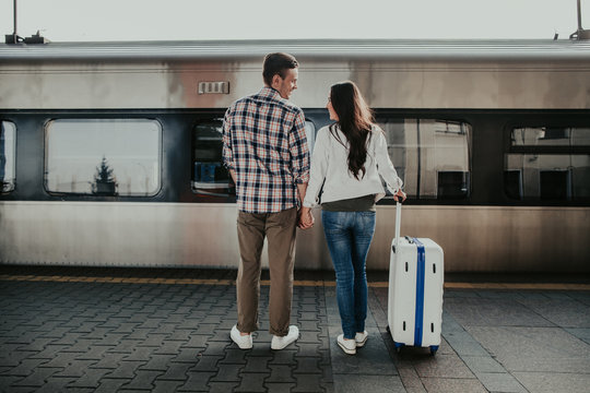 Full length positive man communicating with girl on platform opposite train before trip. She holding baggage in hand and turning back to camera