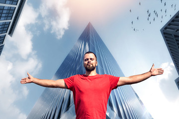 Fototapeta na wymiar Portrait of free and happy man with both arms wide open facing the bright sunshine against blue sky and modern buildings in the background/Young man stretching his arms over blue sky background
