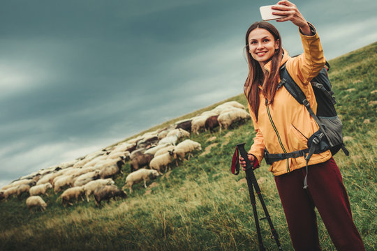 Joyful yougn brunette woman standing near herd of sheep while making selfies on the hills