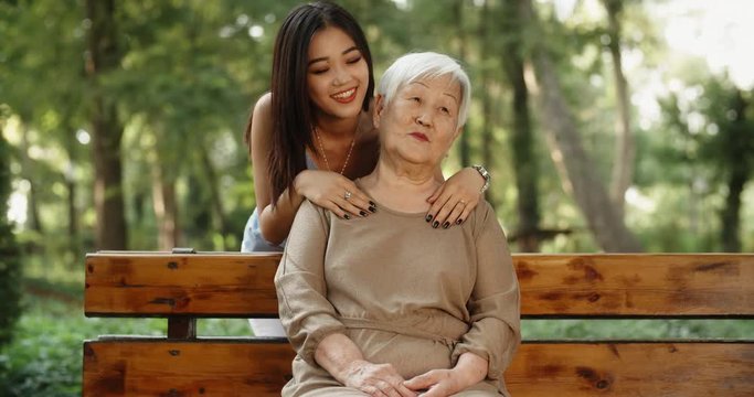 Teen asian granddaughter hugging and kissing her happy mature grandmother sitting on bench, smiling. 4k