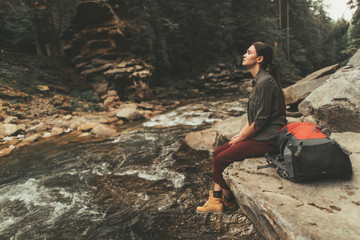 Delighted beautiful young woman sitting on the bank of mountain river while enjoying amazing scenary