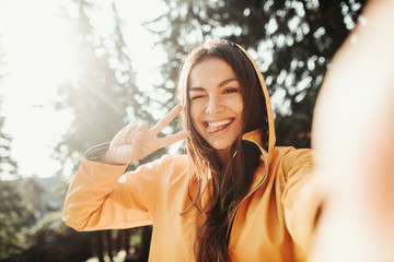 Be positive. Close up portrait of cute girl in yellow jacket showing peace gesture while taking...