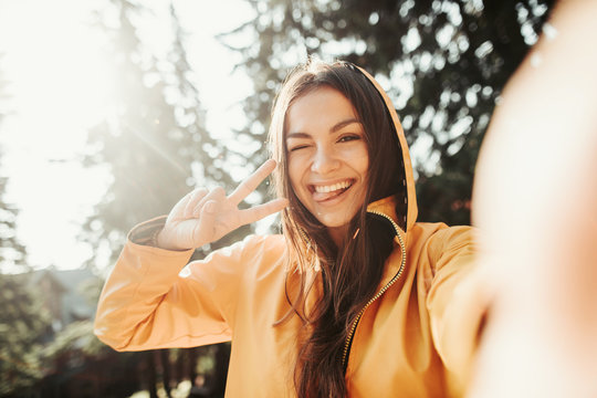 Be positive. Close up portrait of cute girl in yellow jacket showing peace gesture while taking photo with smartphone. She is winking and sticking her tongue out