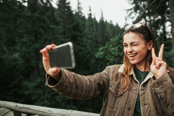 Having fun in the woods. Portrait of charming young lady showing peace gesture while taking photo...