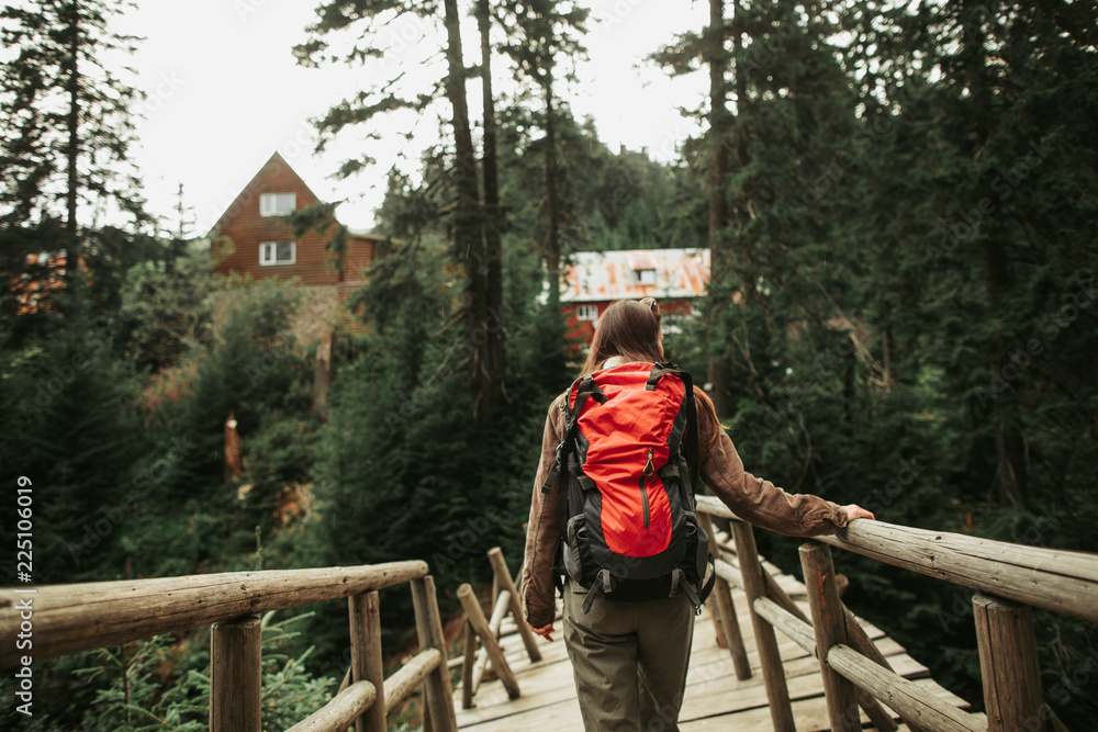 Wall mural one more quest. back view of brave girl with backpack crossing old footbridge in coniferous forest. 