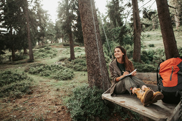 Fototapeta premium I like this place. Portrait of charming young lady holding notebook with pen while looking away with smile. Trees and green plants on background
