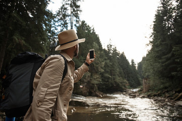 Taking photos. Active young traveler wearing a big hat and carrying heavy backpack while holding his modern smartphone and taking photos of the beautiful river