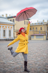 A red-haired woman in a yellow coat and a red beret is jumping with a red umbrella. A smiling beautiful woman in striped pants and a warm jacket flies with an umbrella, like Merry Poppins.