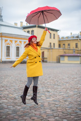 A red-haired woman in a yellow coat and a red beret is jumping with a red umbrella. A smiling beautiful woman in striped pants and a warm jacket flies with an umbrella, like Merry Poppins.