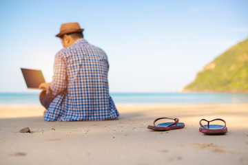 Selective focus,Young Asian man using laptop in blue shirt sitting on the beach,Freelancer working on sea beach background,copy space.