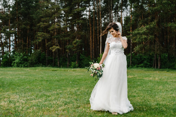 Obraz na płótnie Canvas Young beautiful bride in an elegant dress is standing on the field near the forest and holding bouquet of pink flowers and greens with ribbon at nature. Outdoors. After the wedding ceremony.