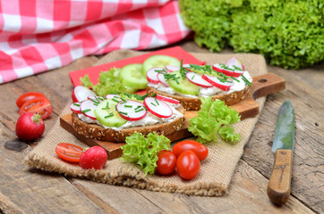 Wholemeal grain bread with curd cheese, fresh radish, cucumber, tomatoes and knife on a wooden...