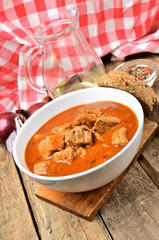 Close-up of pork goulash with pieces of meat in a bowl, garlic, pepper, onion, jug with oil and red checkered tablecloth in the background - vertical photo