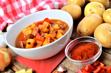 Potato and sausage goulash in dish, pepper, onion and garlic on wooden table
