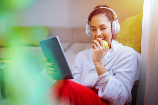 Cute young girl listening to the music and looking at the tablet. Eating green apple and relaxing.