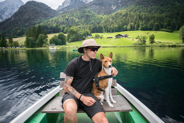 Young man in hiking or camping outfit sits together with his puppy pet, basenji breed dog on rowing boat, swimming in alpine mountain lake, enjoys healthy active lifestyle