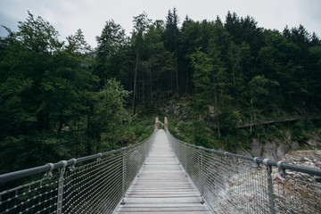 Fototapeta na wymiar View of moody and authentic wooden hanging bridge in lush green forest in mountains over fast river, concept exploration, camping and outdoor activities. New horizons and adventures lifestyle