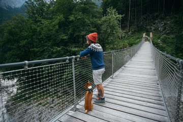 Trendy and hip, young man in adventure gear, hiking or camping wool blue knit sweater and orange beanie, with brown basenji dog or puppy stands on hanging wooden bridge over mountain forest river