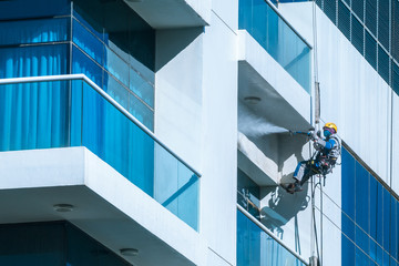 Worker wearing safety harness washes glass facade at height on modern high rise building. Professional rope access