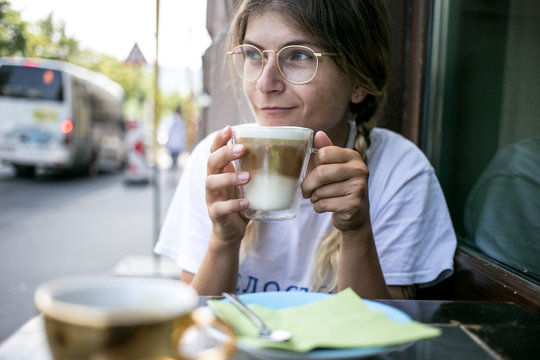 Cute millennial young pretty woman with braids relaxed and chill sits in cafe terrace outside, enjoys coffee drink during summer lunch break, drinks latte layered cappuccino from glass cup
