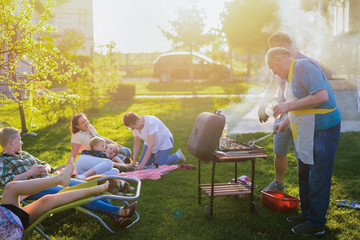 Big happy family sitting in a backyard and enjoying while grandfather is making barbeque for them. Summer day in nature.