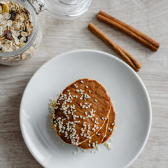 Still life morning breakfast of pancakes with honey and sprinkled with sesame seeds, cinnamon sticks and muesli in glass on a white plate on a wooden background.