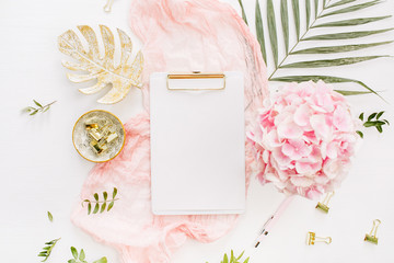 Modern home office desk workspace with blank paper clipboard, pink hydrangea flowers bouquet, tropical palm leaf, pastel blanket, monstera leaf plate and accessories on white background. Flatlay.