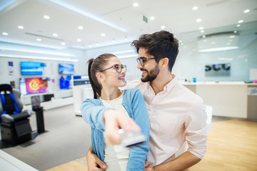 Young happy couple looking for TV in electronic shore . Girl is holding remote controller and looking at her boyfriend.