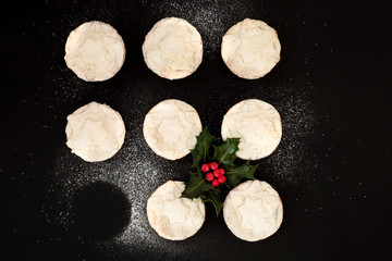 Fototapeta na wymiar Delicious freshly baked Christmas mince pies with one missing, with holly berry leaf sprig and icing sugar dusting on black background. Top view.