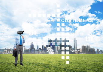 Camera headed man standing on green grass against modern cityscape