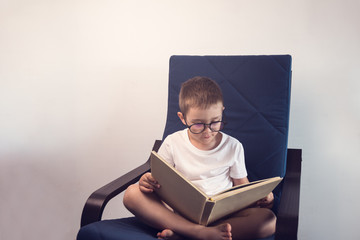 Obraz premium Young kid reading an interesting book sitting on the blue armchair. Concept of reading an interesting and exciting book everywhere.