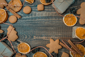 Christmas top view over a brown wooden background with gift boxes packed in a craft paper, cookies, dryed oranges, pine cones, craft ropes and cinnamon sticks placed in circle. Flat lay, copy space, h