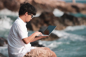 Relaxed young Asian man using laptop at sea shore. Internet of things concept.