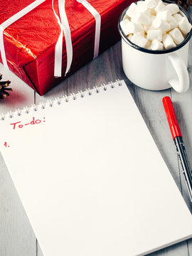 Christmas to-do list. Xmas gifts shopping planning. Make shopping or to-do list for Christmas. Notebook, mug hot chocolate with marshmallows and New Year's gift on gray wooden background.
